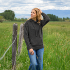 A woman wearing a dark charcoal gray knit hoodie and blue jeans stands in a grassy field with a rustic wooden fence and mountains in the background.