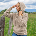 A woman wearing a beige speckled knit hooded sweater leans against a rustic wooden fence post in an outdoor grassy field under a cloudy sky.