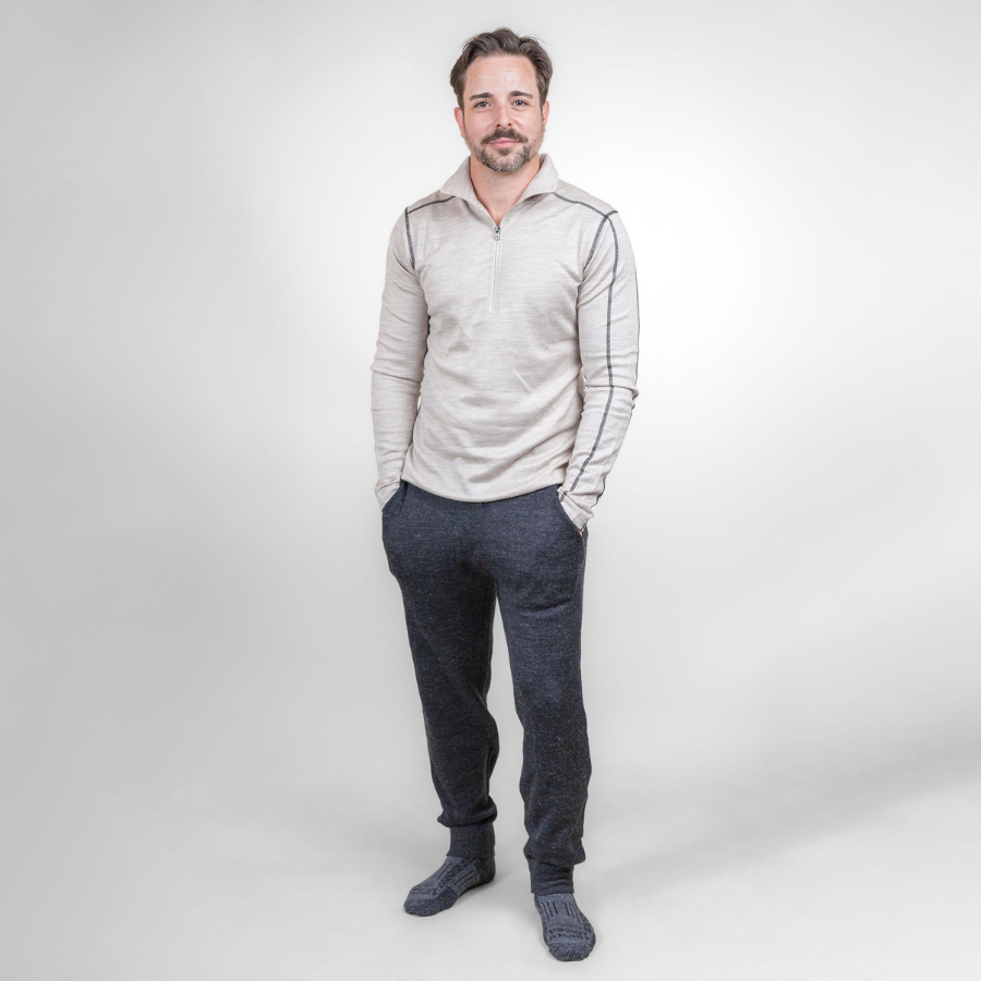 A smiling man standing with his hands in his pockets in front of a white background. He is wearing gray Alpacas of Montana adventure hiking socks, oatmeal Alpacas of Montana men's base layer top, and a pair of charcoal gray Alpacas of Montana soft cozy comfortable lightweight lounge moisture wicking antimicrobial exercise men's joggers sweatpants made from alpaca wool for lounging, comfort, exercise, hiking, skiing
