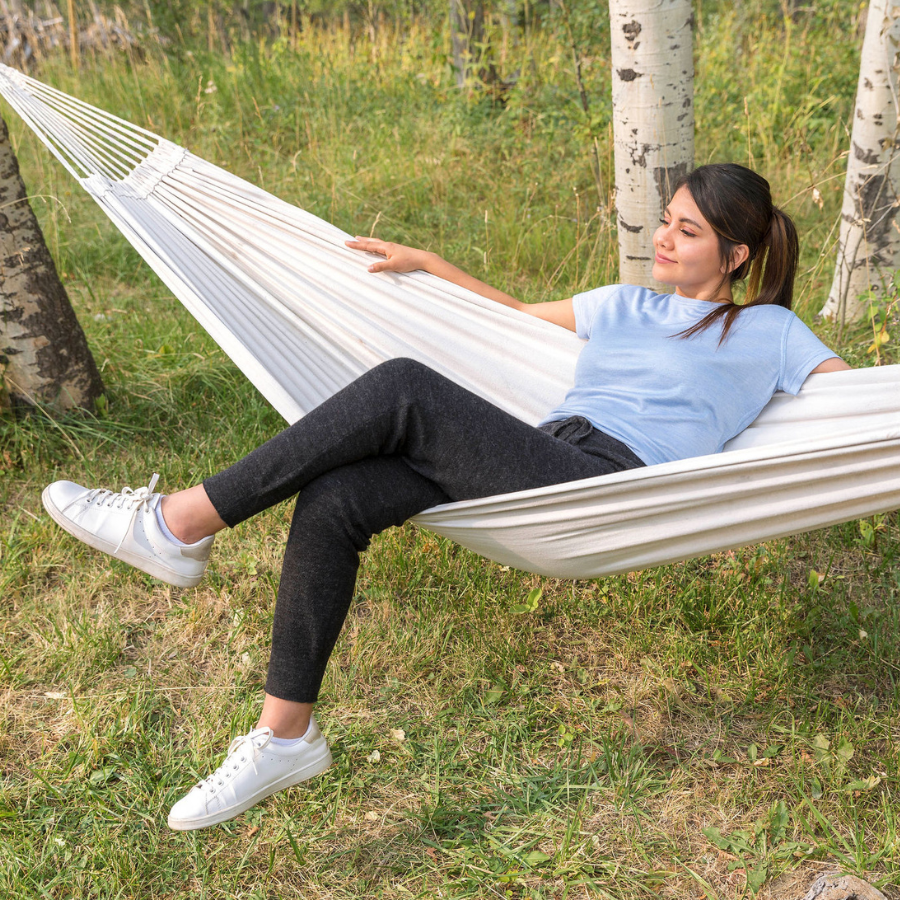 A smiling brown haired woman lounging in a hammock in a grassy forest wearing white sneakers, a light blue tee shirt, and A brown haired woman standing against a white background. She is wearing white sneakers, a white tank top, and charcoal gray Alpacas of Montana soft cozy comfortable lightweight lounge moisture wicking antimicrobial exercise women's 24/7 boyfriend joggers sweatpants made from alpaca wool for lounging, comfort, exercise, hiking, skiing