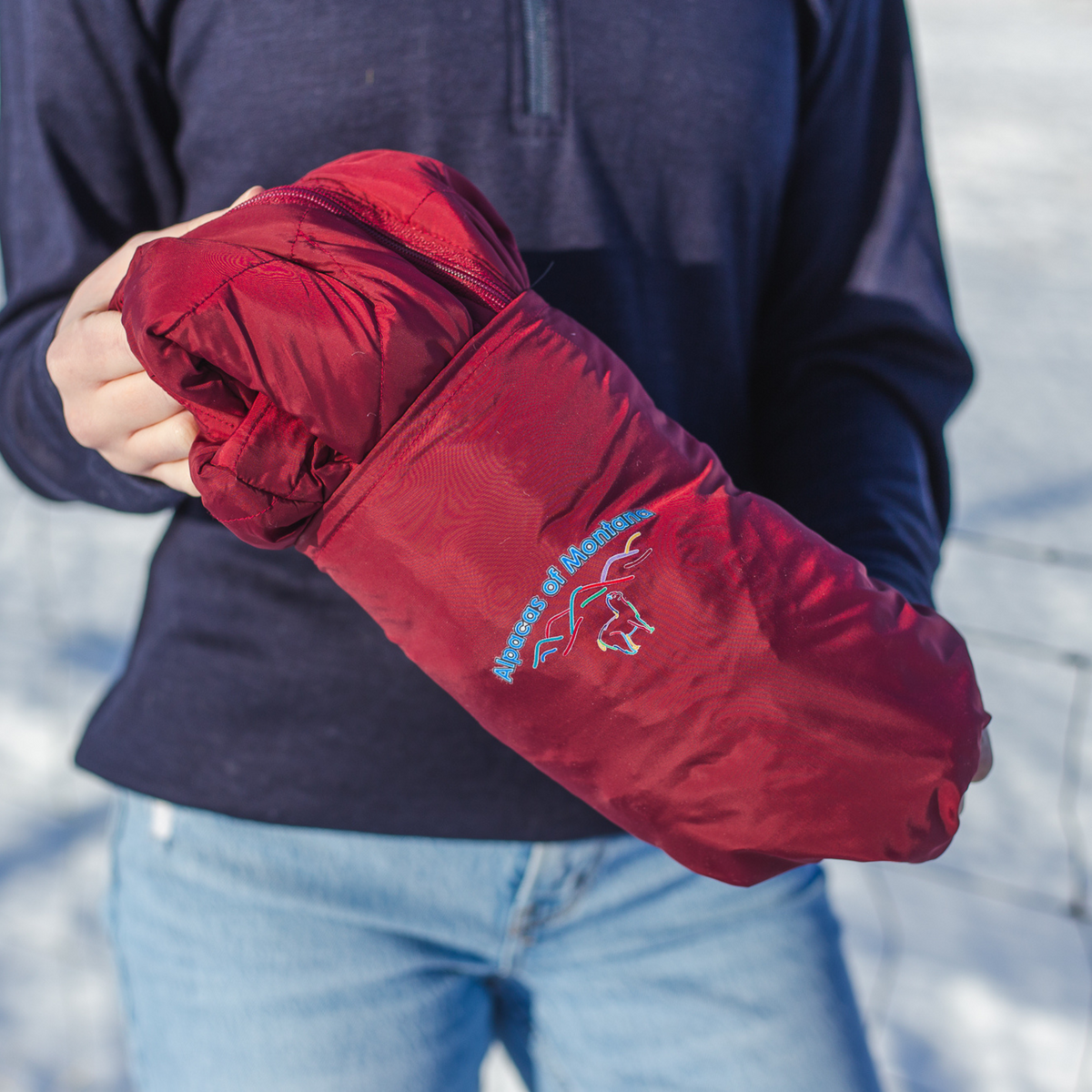 A person holding a red fabric bag with the Alpacas of Montana logo on it. Inside the bag is a cranberry red Alpacas of Montana warm top layer insulated cozy comfortable thermal winter outerwear lightweight alpaca wool insulated women&#39;s tempest coat jacket parka for skiing, snowboarding, ice fishing, hunting, camping, arctic, travel, outdoors