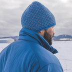 A man with a black beard facing away from the camera in a snowy scene. He is wearing a blue Alpacas of Montana men's tempest lite jacket and a blue and gray Alpacas of Montana extremely warm cozy soft windproof comfortable antimicrobial moisture wicking thermal alpaca fleece wool winter fleece lined beanie hat for hiking, skiing, snowshoeing, sledding, outdoors.