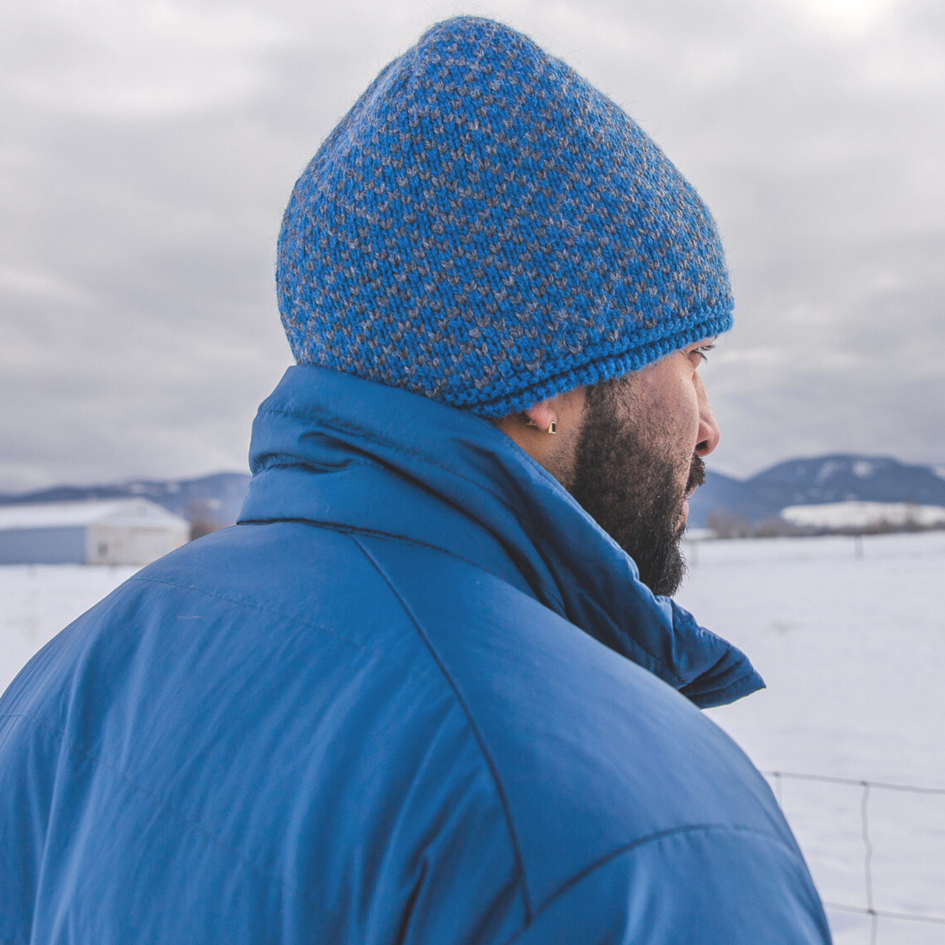 A man with a black beard facing away from the camera in a snowy scene. He is wearing a blue Alpacas of Montana men's tempest lite jacket and a blue and gray Alpacas of Montana extremely warm cozy soft windproof comfortable antimicrobial moisture wicking thermal alpaca fleece wool winter fleece lined beanie hat for hiking, skiing, snowshoeing, sledding, outdoors.