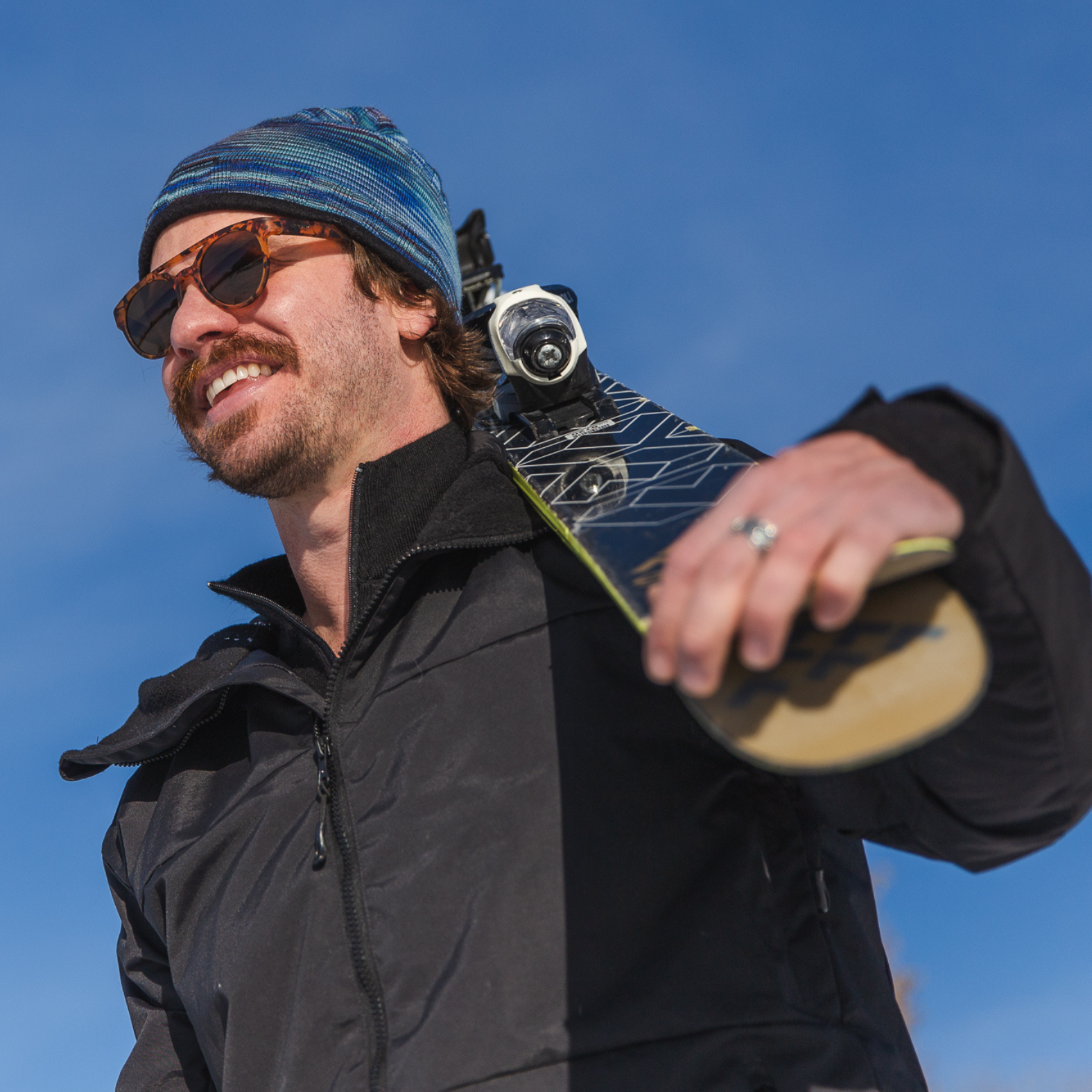 A man holding a pair of skis with blue sky in the background. He is wearing the Alpacas of Montana graphite color Granite Peak Parka and a multi-blue, gray, black, green, purple, and white outerwear hiking skiing winter warm soft comfortable Backcountry beanie