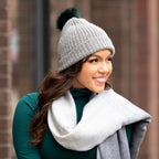 A brown-haired woman smiling wearing a green shirt, a gray Alpacas of Montana shawl, and a soft warm winter cozy moisture wicking comfortable fashionable light gray alpaca wool beartooth beanie with dark forest green pom pom
