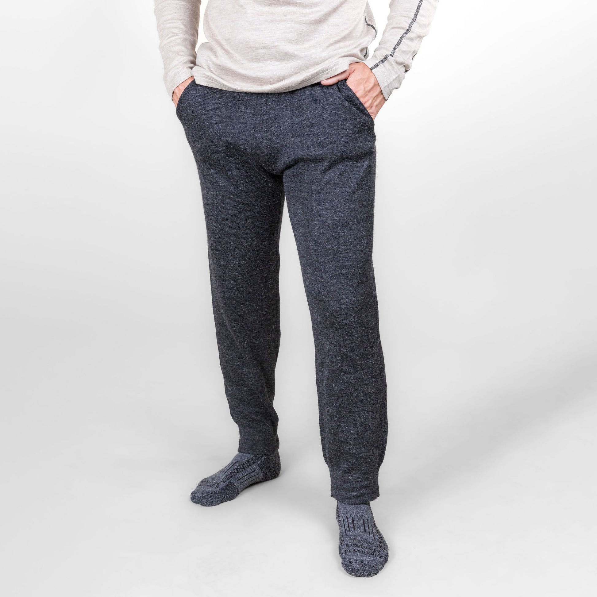 A man standing in front of a white background wearing an oatmeal Alpacas of Montana men's base layer top, gray Alpacas of Montana adventure hiking socks, and charcoal gray Alpacas of Montana soft cozy comfortable lightweight lounge moisture wicking antimicrobial exercise men's joggers sweatpants made from alpaca wool for lounging, comfort, exercise, hiking, skiing