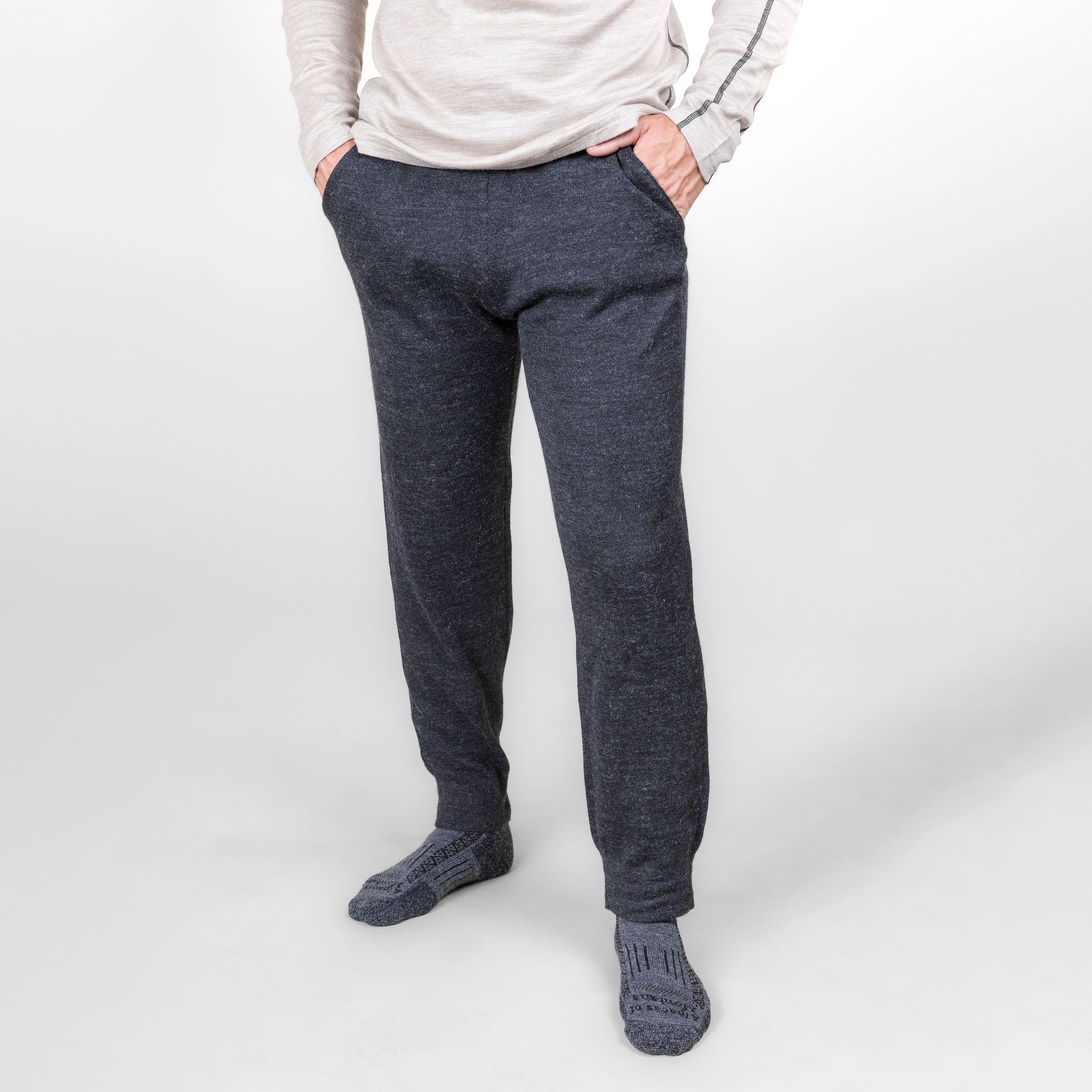 A man standing in front of a white background wearing an oatmeal Alpacas of Montana men's base layer top, gray Alpacas of Montana adventure hiking socks, and charcoal gray Alpacas of Montana soft cozy comfortable lightweight lounge moisture wicking antimicrobial exercise men's joggers sweatpants made from alpaca wool for lounging, comfort, exercise, hiking, skiing