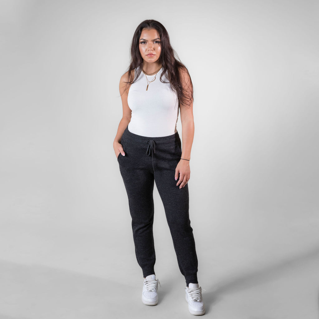 A brown haired woman standing against a white background. She is wearing white sneakers, a white tank top, and charcoal gray Alpacas of Montana soft cozy comfortable lightweight lounge moisture wicking antimicrobial exercise women's 24/7 boyfriend joggers sweatpants made from alpaca wool for lounging, comfort, exercise, hiking, skiing