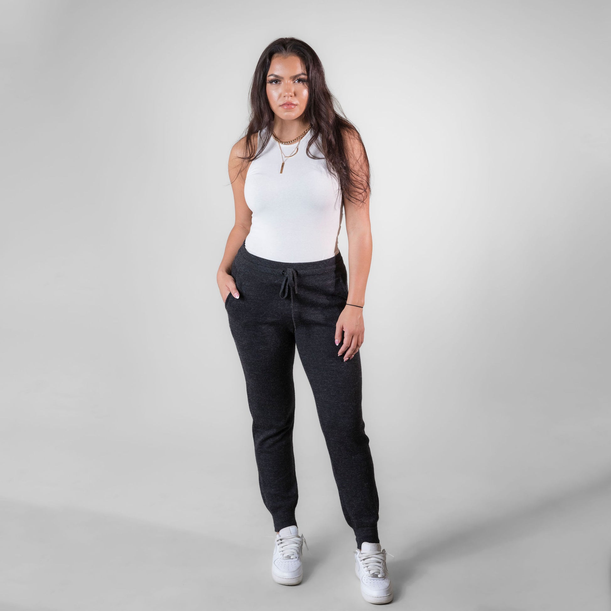 A brown haired woman standing against a white background. She is wearing white sneakers, a white tank top, and charcoal gray Alpacas of Montana soft cozy comfortable lightweight lounge moisture wicking antimicrobial exercise women's 24/7 boyfriend joggers sweatpants made from alpaca wool for lounging, comfort, exercise, hiking, skiing