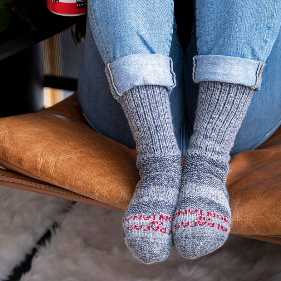 A close up photo of a pair of feet, the person is wearing blue jeans and sitting with their feet pulled up onto a brown leather chair. They are wearing Alpacas of Montana cozy soft warm comfortable thermal moisture wicking everyday winter fishing hiking snowshoeing hunting outdoors silver gray extra cushion boot socks.