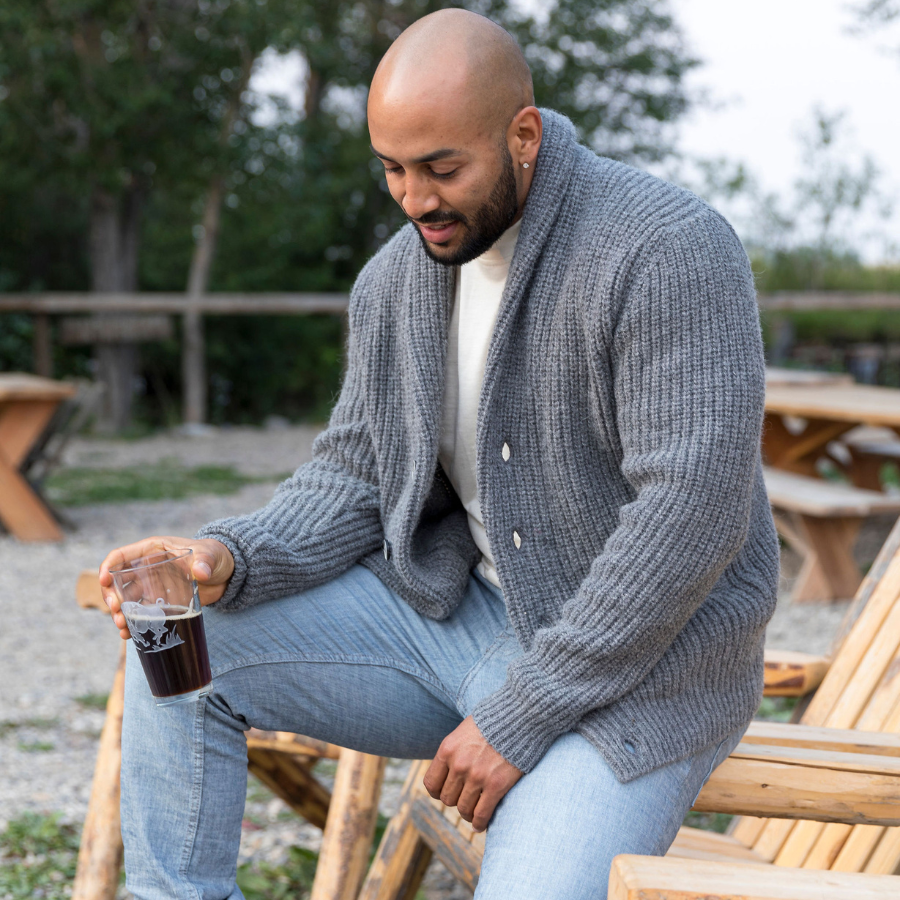 A bald man with a black beard looking at the ground while sitting on a wooden lounge chair holding a glass with a drink in it. He is wearing light blue jeans, a white shirt, and an Alpacas of Montana cozy comfortable soft moisture wicking formal casual stylish upcycled men's medium gray button up chunky knit ribbed cardigan.