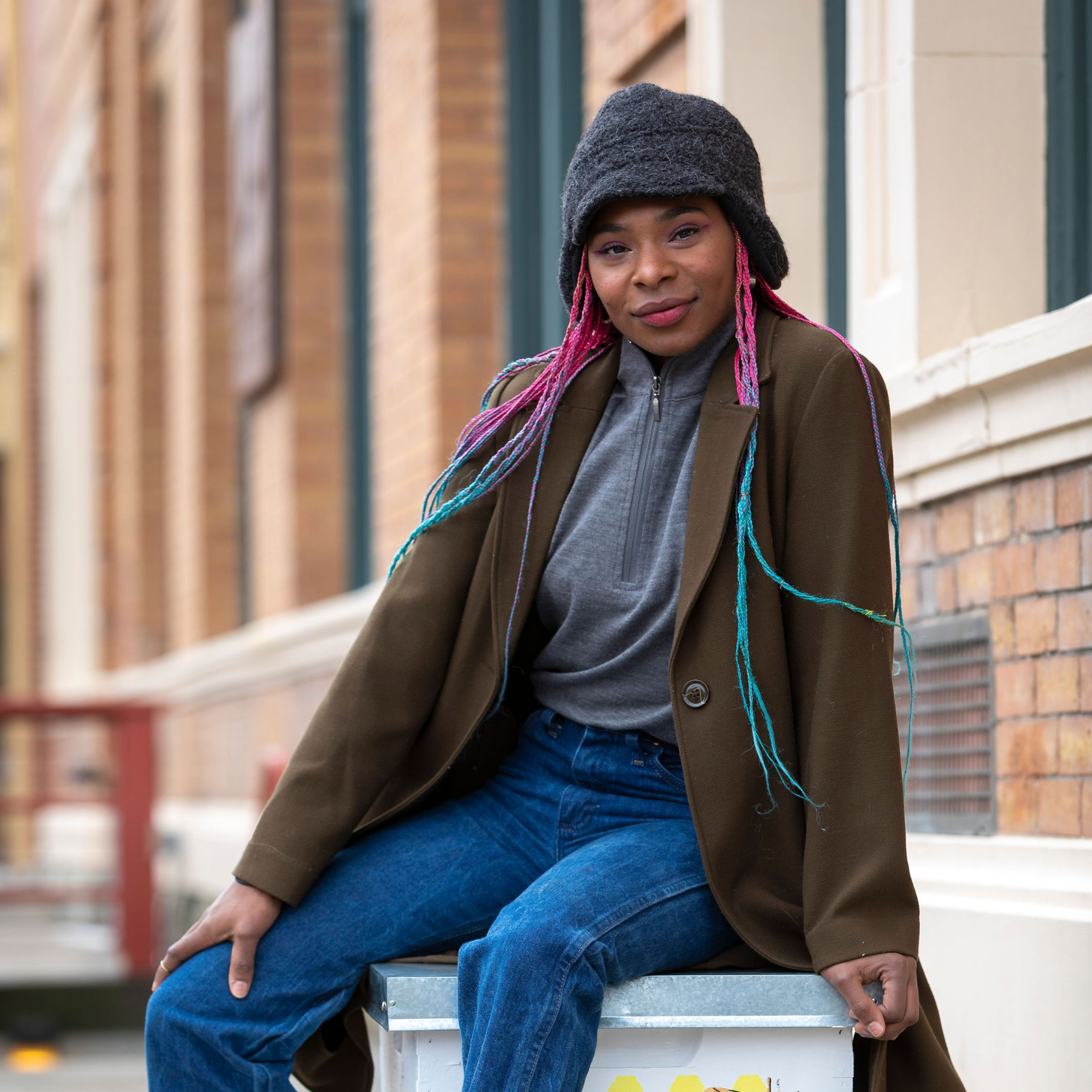 A sitting woman with long pink, purple, and blue hair smiling and looking at the camera. She is wearing blue jeans, a gray Alpacas of Montana women's base layer top, a long brown jacket, and a dark gray extremely warm cozy soft windproof comfortable moisture wicking thermal winter alpaca fleece wool city commuter hat.