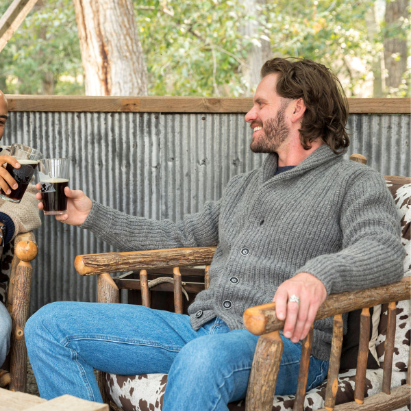 A man sitting in a wooden lounge chair outdoors smiling and looking to the left while he clinks drink glasses with another man. He is wearing blue jeans and an Alpacas of Montana cozy comfortable soft moisture wicking formal casual stylish upcycled men's medium gray button up chunky knit ribbed cardigan.
