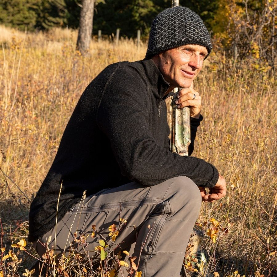 A man kneeling in a golden grassy field looking off into the distance. He is wearing gray pants, a black Alpacas of Montana men's mid-layer quarter zip top, and a black and gray Alpacas of Montana extremely warm cozy soft windproof comfortable antimicrobial moisture wicking thermal alpaca fleece wool winter fleece lined beanie hat for hiking, skiing, snowshoeing, sledding, outdoors, hunting.