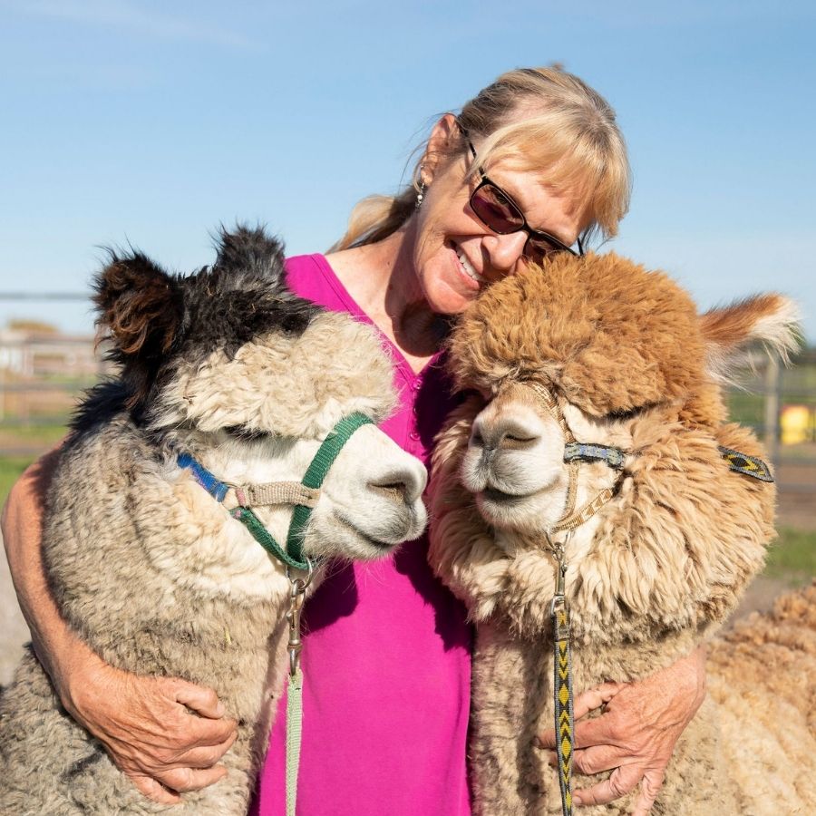 woman smiling with her arms around two alpacas hugging them