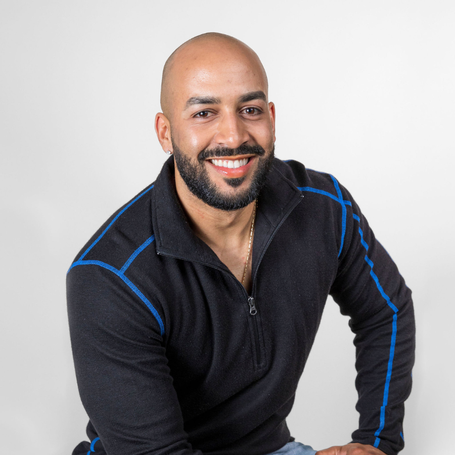 A smiling bald man with a black beard sitting in front of a white background. He is wearing a black with blue stitching Alpacas of Montana warm thermal soft cozy comfortable activewear outerwear athletic moisture wicking antimicrobial men's fashion stylish luxury mid-layer quarter-zip alpaca wool long sleeve pullover top for outdoors camping climbing hiking skiing hunting fishing running winter.