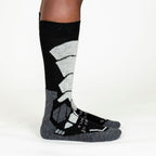 A person's lower legs standing sideways with a white background wearing light gray, dark grey, and black Alpacas of Montana cozy comfortable soft warm thermal winter freezing temperatures antimicrobial moisture wicking alpaca wool ski and snowboard socks for snowshoeing, skiing, snowboarding, ice fishing, outdoors.