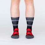 A photo of a person's lower legs standing against a white background wearing black, scarlet red, multi-gray, and white accent Alpacas of Montana soft cozy comfortable activewear outerwear athletic workout moisture wicking antimicrobial cushioned light compression engineered high-tech mid-crew hiking sock for walking, skiing, climbing, hunting, camping, fishing, exercise, biking