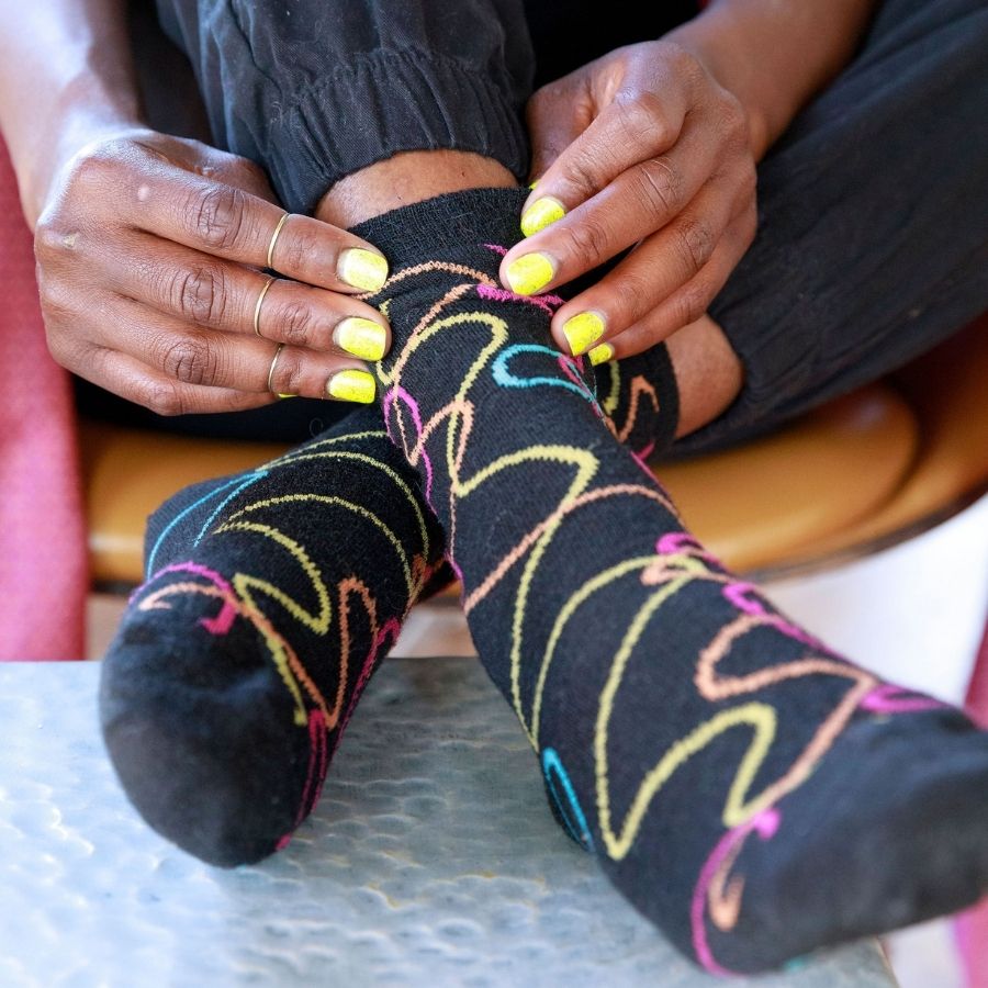A close up photo of a pair of hands with painted yellow nails pulling up a pair of the Alpacas of Montana colorful yellow, orange, pink, and blue squiggle pattern black casual lounge fashion comfortable soft cozy everyday moisture wicking alpaca wool Night Life socks.