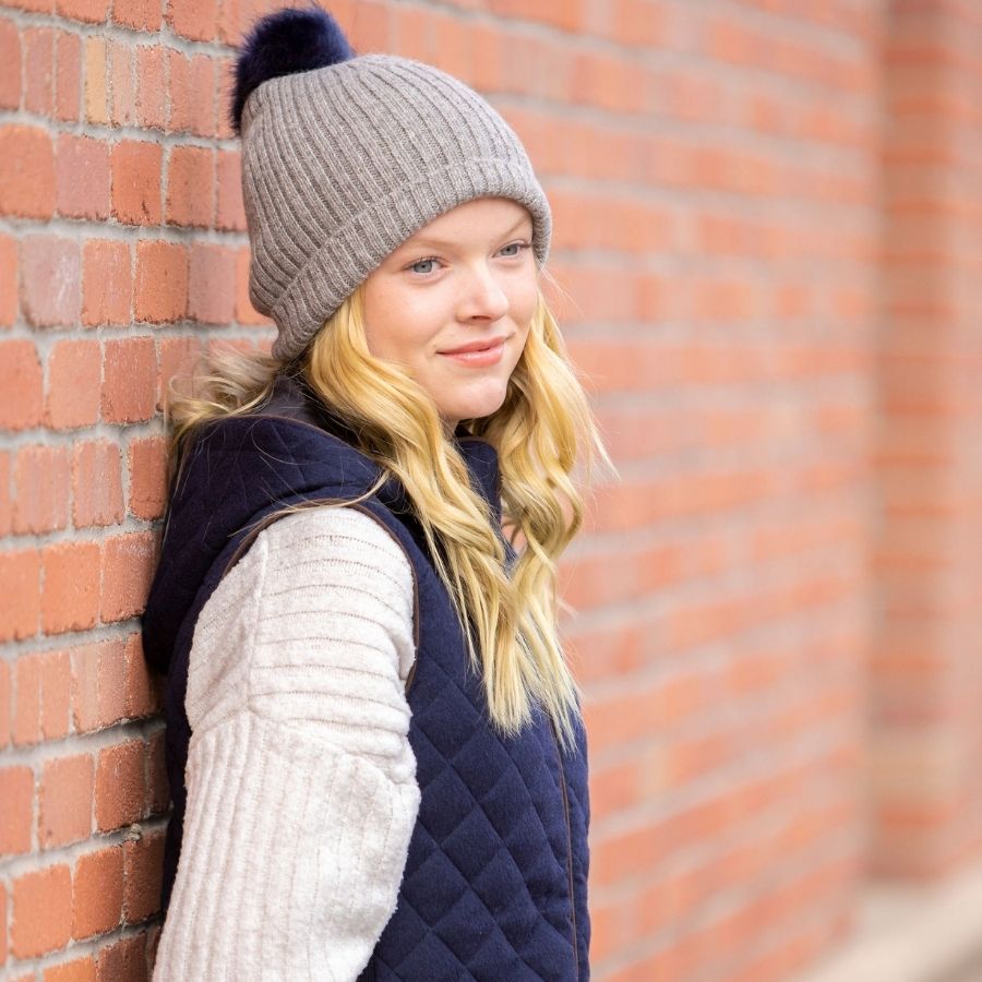 woman looking off into the distance leaned against a brick building wearing gray ribbed alpaca wool beartoothe beanie with blue pom pom 