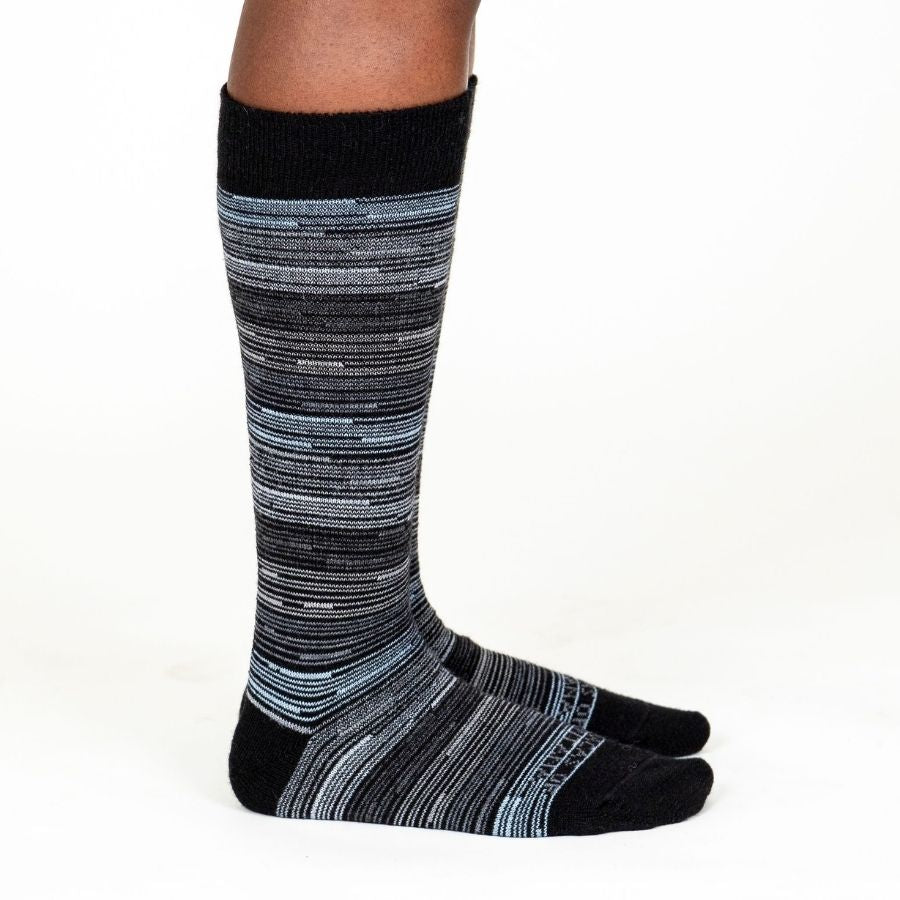 A person's lower legs standing sideways with a white background wearing Alpacas of Montana black, gray, and white casual lounge fashion comfortable soft cozy everyday moisture wicking alpaca wool striped socks.