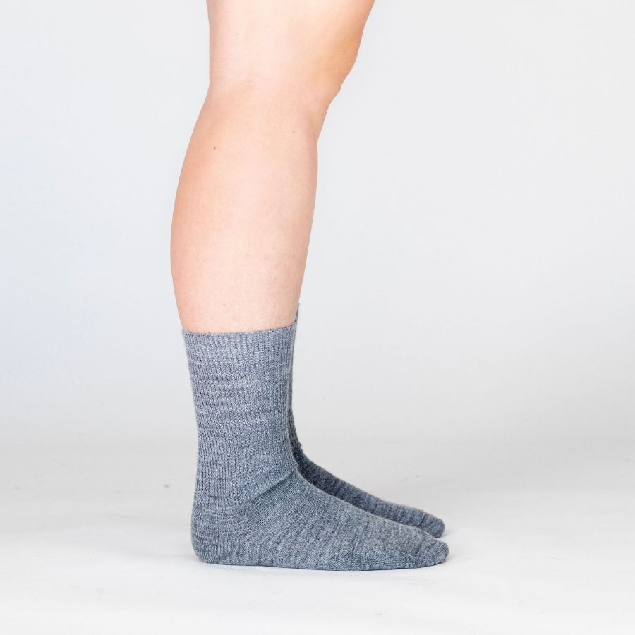 Side view of a person&#39;s lower legs against a white background wearing medium light gray Alpacas of Montana professional soft comfortable moisture wicking business casual alpaca wool dress socks for men and women.