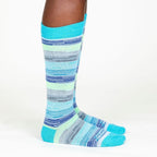 A person's lower legs standing sideways with a white background wearing Alpacas of Montana colorful navy, cobalt, sky blue, lime green, gray, and black casual lounge fashion comfortable soft cozy everyday moisture wicking alpaca wool striped socks.