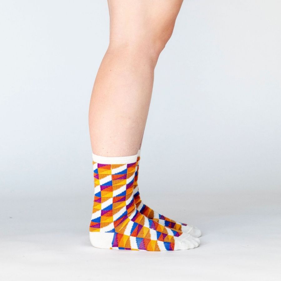 A person's lower legs standing sideways wearing a pair of Alpacas of Montana white, yellow, gold, orange, purple, and blue triangle pattern casual lounge fashion comfortable soft cozy everyday moisture wicking alpaca wool Swag socks.
