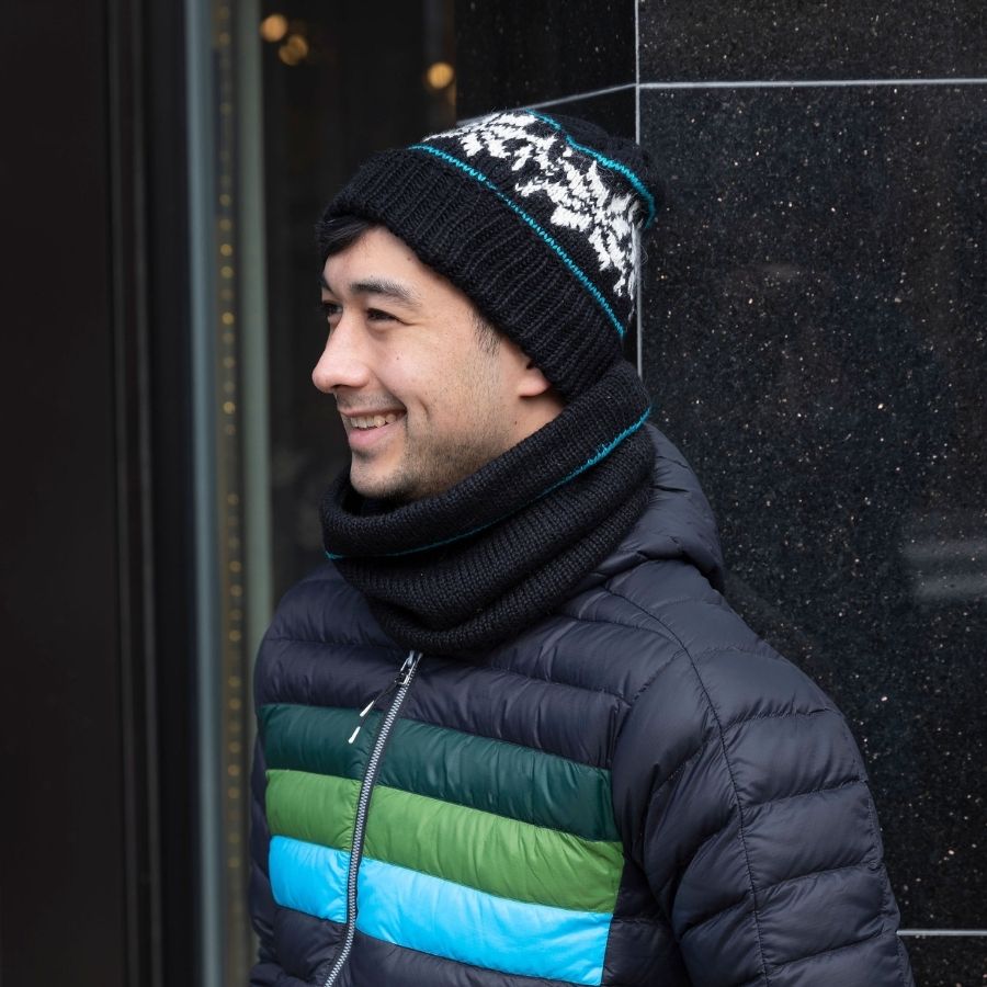 man standing leaning against a wall wearing handmade black alpaca wool cowl and beanie hat with two teal stripes and white snowflakes