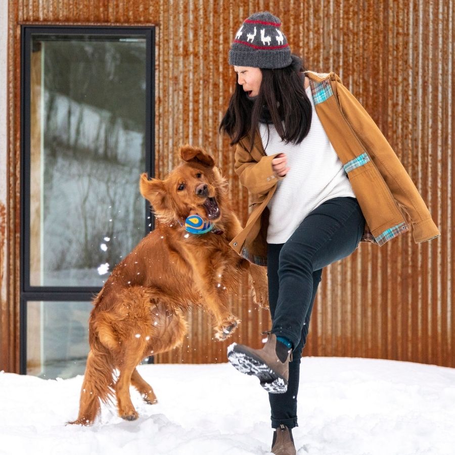 woman kicking snow and looking at golden retriever dog jumping for ball while wearing gray alpaca wool beanie with two red stripes and white alpacas