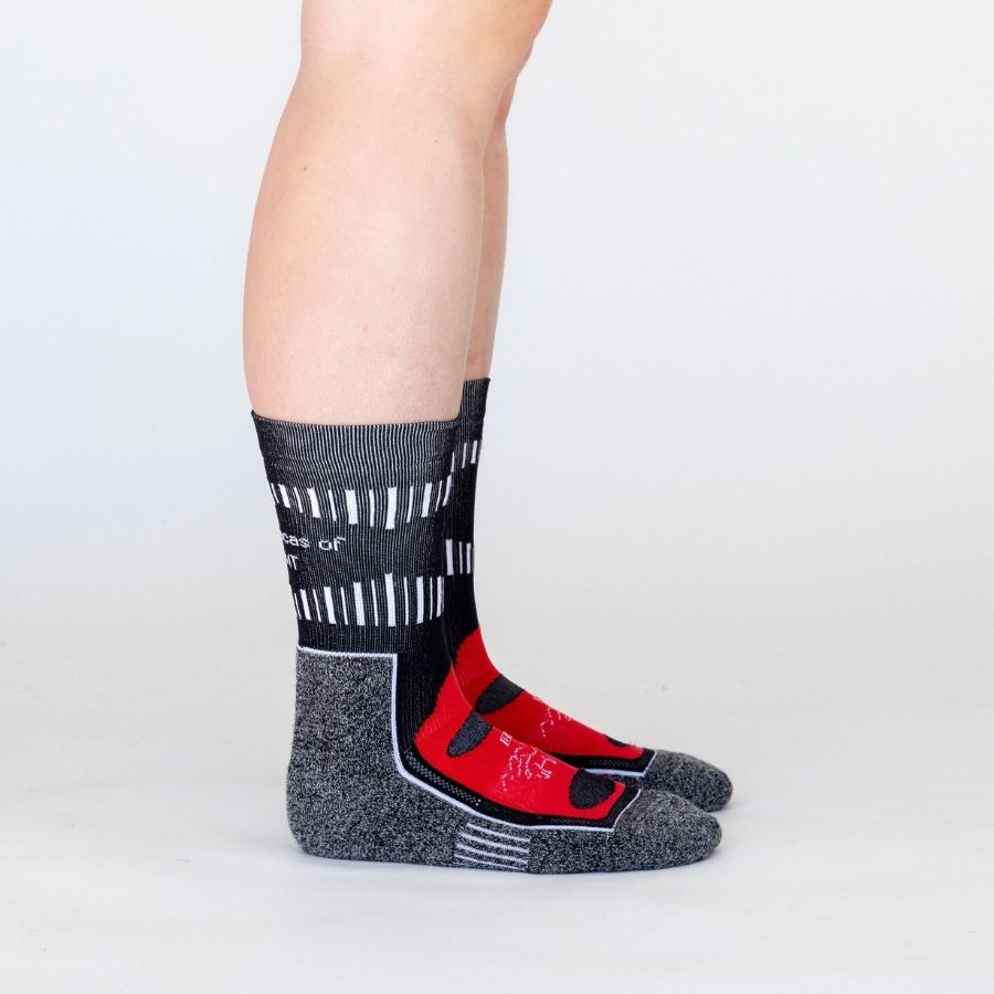 A person standing sideways to the camera against a white background wearing black, scarlet red, and multi-gray Alpacas of Montana soft cozy comfortable activewear outerwear athletic workout moisture wicking antimicrobial cushioned light compression engineered high-tech mid-crew hiking sock for walking, skiing, climbing, hunting, camping, fishing, exercise, biking