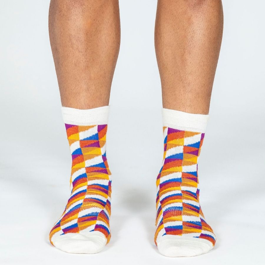 A person's lower legs standing against a white background wearing a pair of Alpacas of Montana white, yellow, gold, orange, purple, and blue triangle pattern casual lounge fashion comfortable soft cozy everyday moisture wicking alpaca wool Swag socks.
