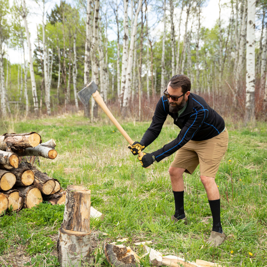 A man with brown hair and a beard chopping wood in a grassy forest clearing. He is wearing sunglasses, slippers, black Alpacas of Montana adventure hiking socks, khaki shorts, and a black with blue stitching Alpacas of Montana warm thermal soft cozy comfortable activewear outerwear athletic moisture wicking antimicrobial men's fashion stylish luxury mid-layer quarter-zip alpaca wool long sleeve pullover top for outdoors camping climbing hiking skiing hunting fishing running winter.