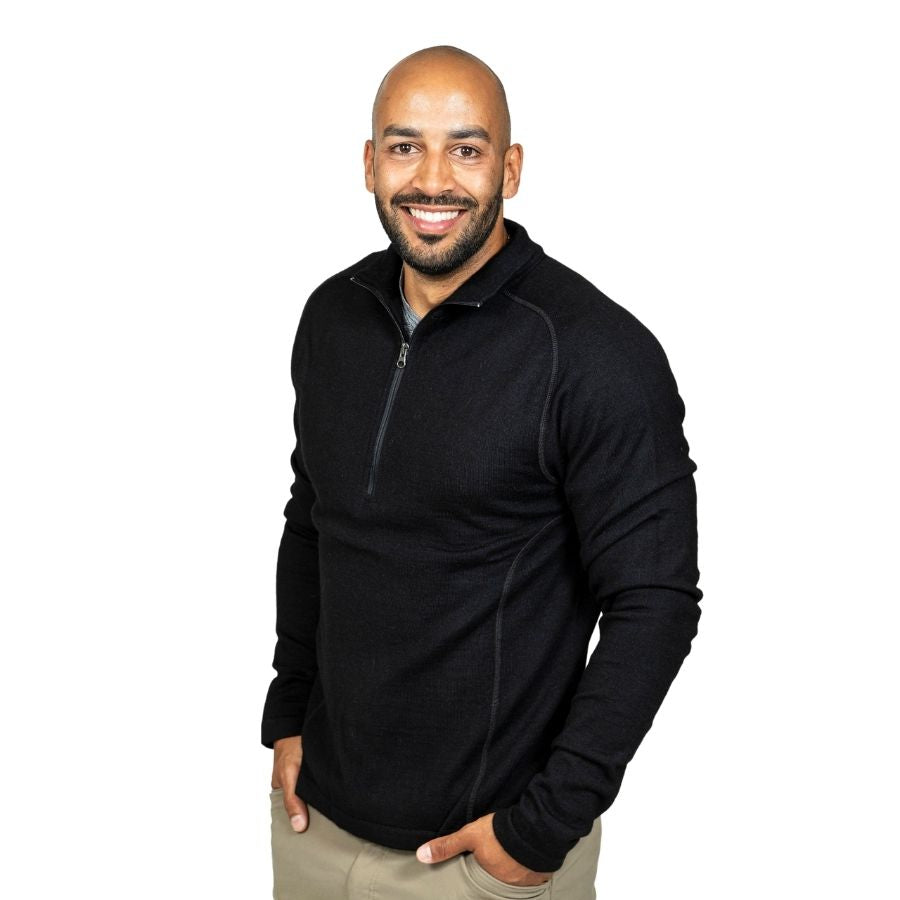 A smiling bald man with a black beard standing in front of a white background. He is wearing khaki pants and a black Alpacas of Montana warm thermal soft cozy comfortable activewear outerwear athletic moisture wicking antimicrobial men's fashion stylish luxury mid-layer quarter-zip alpaca wool long sleeve pullover top for outdoors camping climbing hiking skiing hunting fishing running winter.