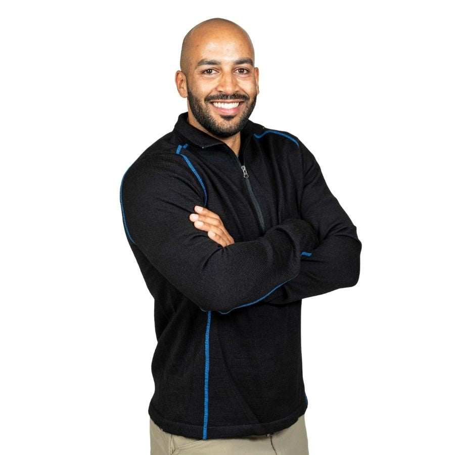 A smiling bald man with a black beard standing with his arms crossed in front of a white background. He is wearing khaki pants and a black with blue stitching Alpacas of Montana warm thermal soft cozy comfortable activewear outerwear athletic moisture wicking antimicrobial men's fashion stylish luxury mid-layer quarter-zip alpaca wool long sleeve pullover top for outdoors camping climbing hiking skiing hunting fishing running winter.