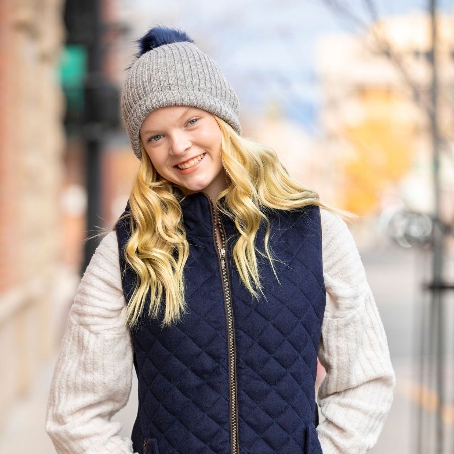 A blonde-haired woman smiling wearing a white sweater, a navy Alpacas of Montana women's hooded vest, and a soft warm winter cozy moisture wicking comfortable fashionable light gray alpaca wool beartooth beanie with dark navy blue pom pom